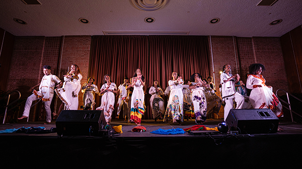 Several student dressed in white traditional African outfits dancing on a stage.