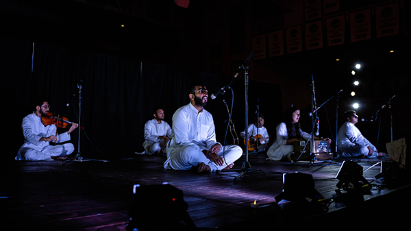 Group of students wearing white garments, sitting on the stage with one students singing into the mic and others playing instruments.
