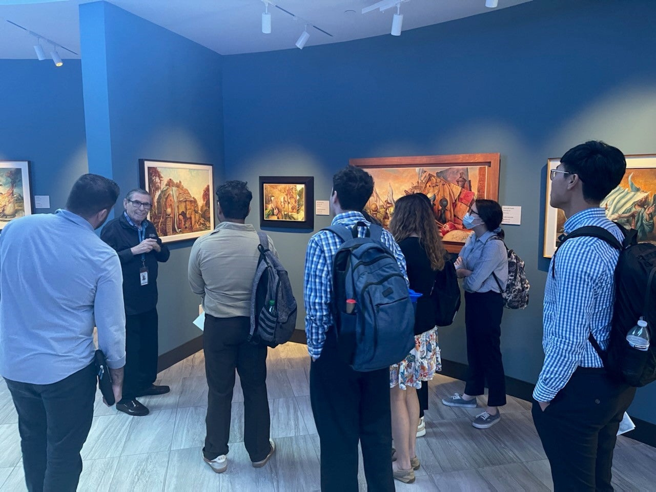 One male standing next to an art piece talking to a group of standing students who are facing him.