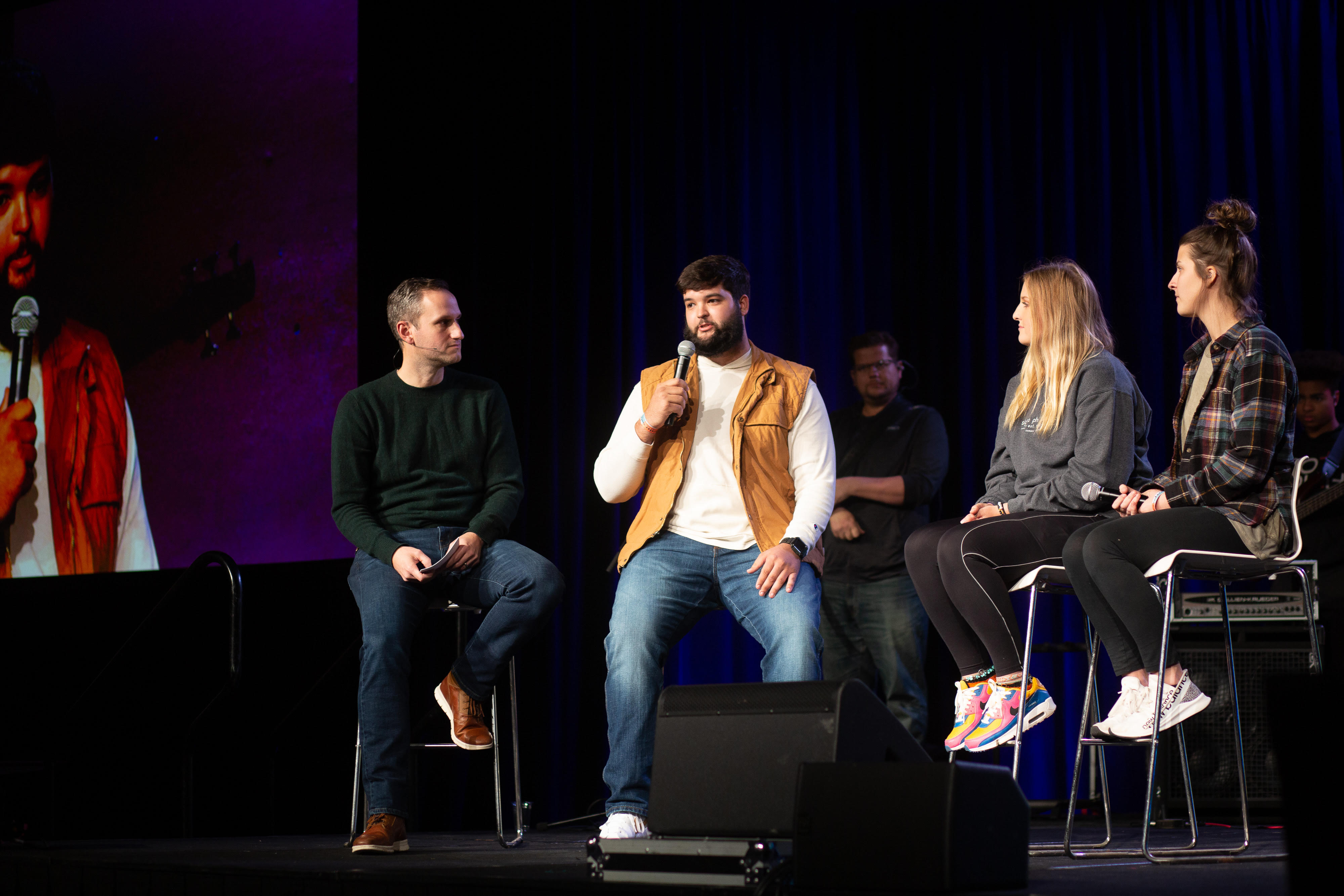 Four people sitting on high chairs on a stage, Connor, who is the second person from left, is holding a mic and speaking into it.