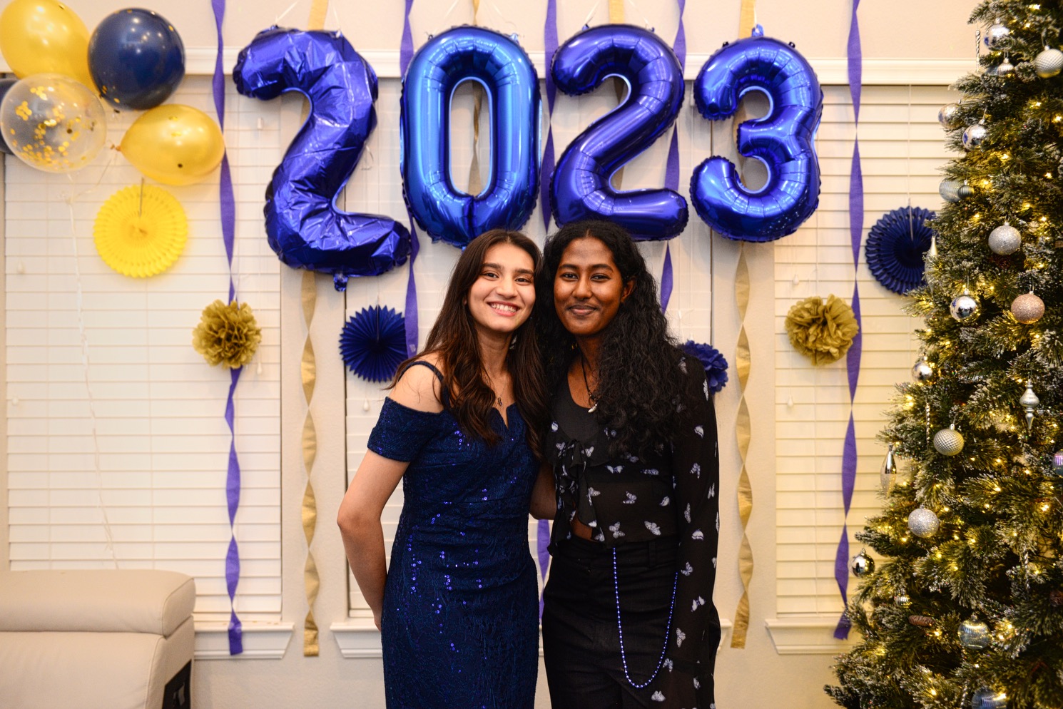 Two females standing next to each other, smiling, with blue balloons that say "2023" and part of a Christmas tree behind them.