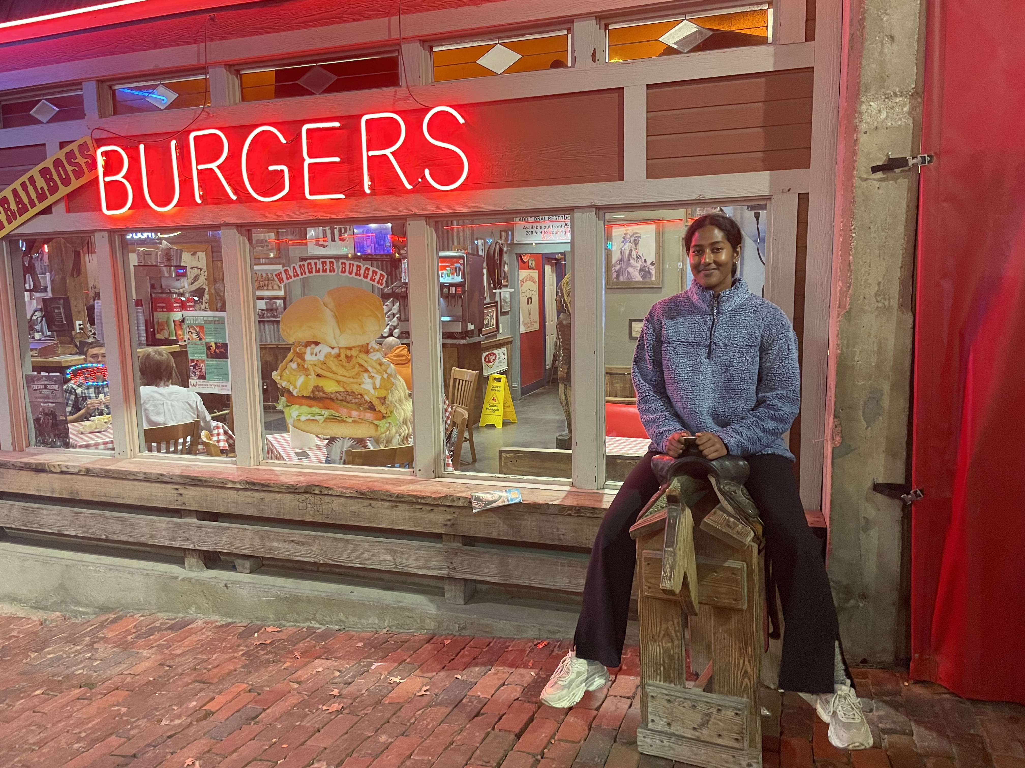 Female sitting on a saddle hoisted on a wooden structure with a "burgers" sign on her right side (left side of image).
