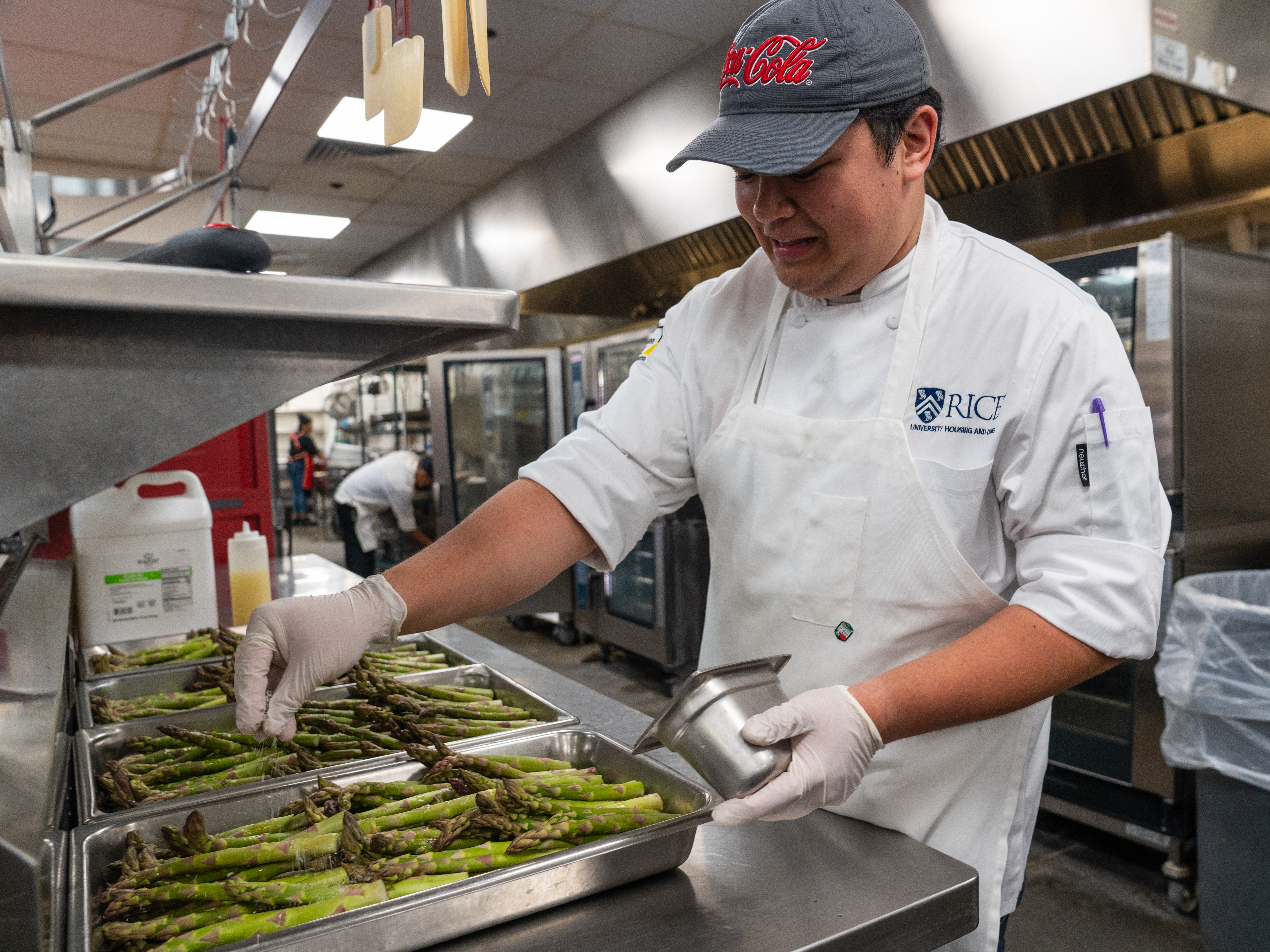 Male wearing a cap, apron, and chef coat salting asparagus in a metal tray.