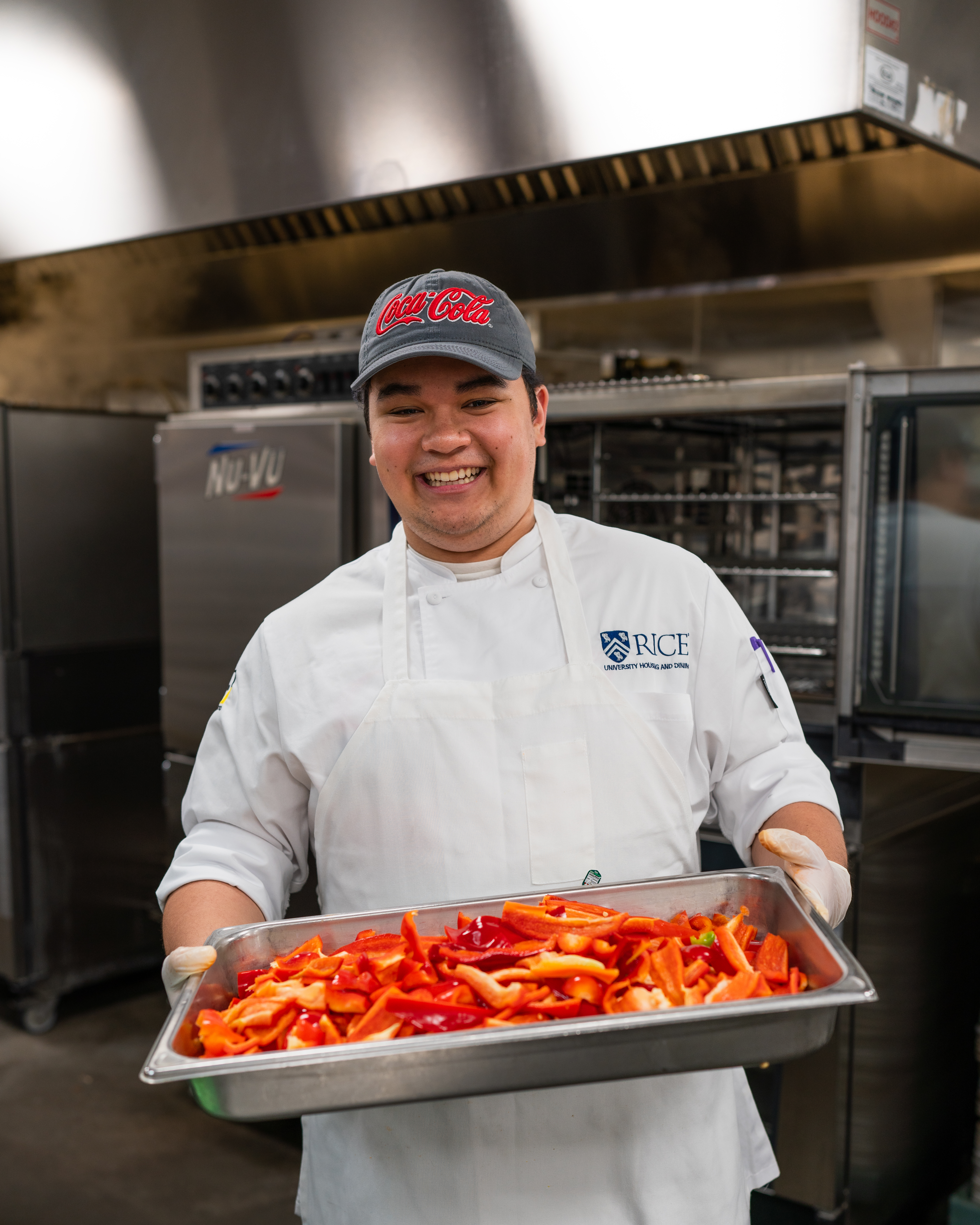 Male wearing an apron and chef coat holding a metal tray of raw, sliced red bell peppers. 