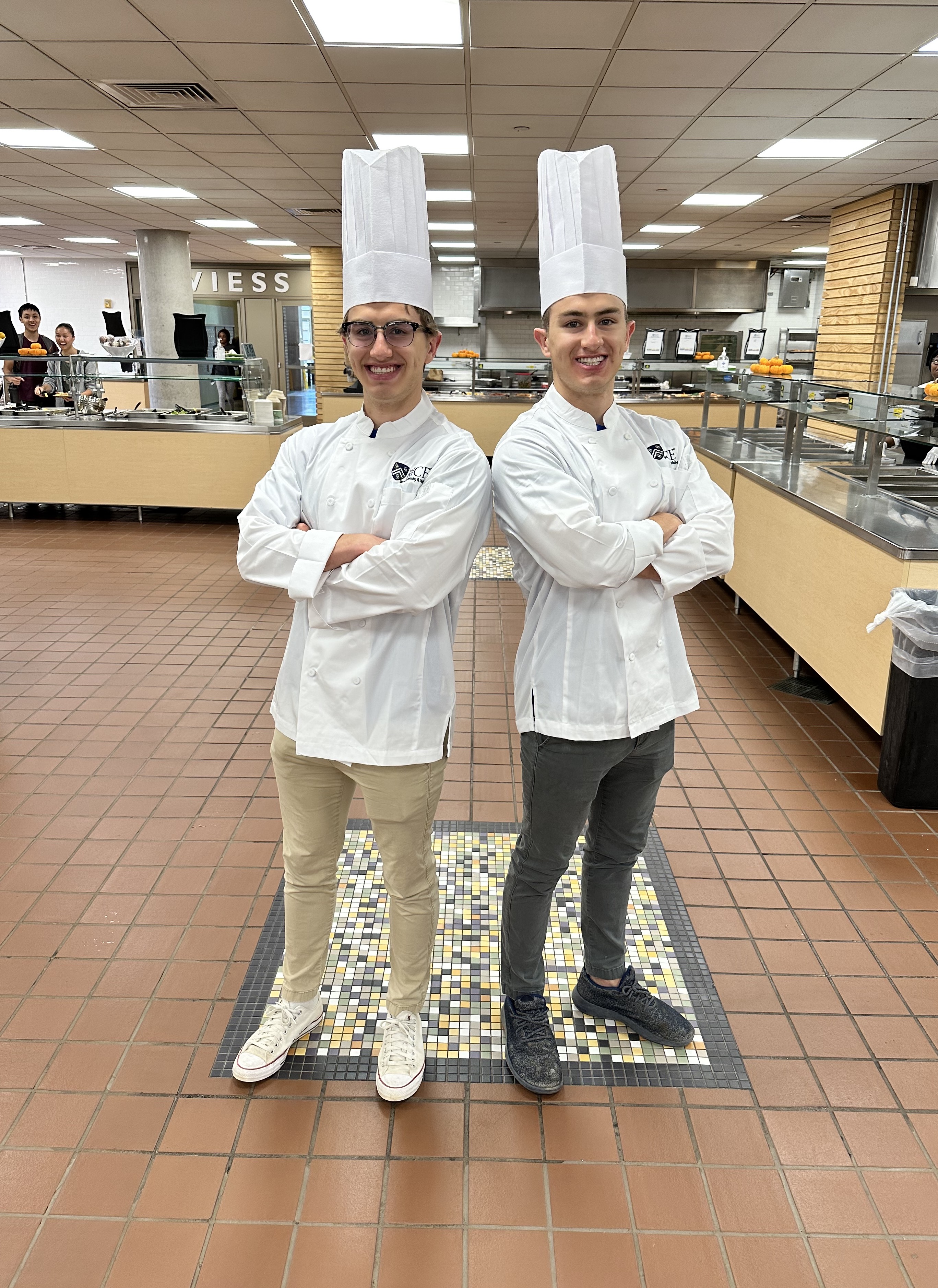 Two male students wearing a chef coat and chef hat with arms crossed and backs against each other.