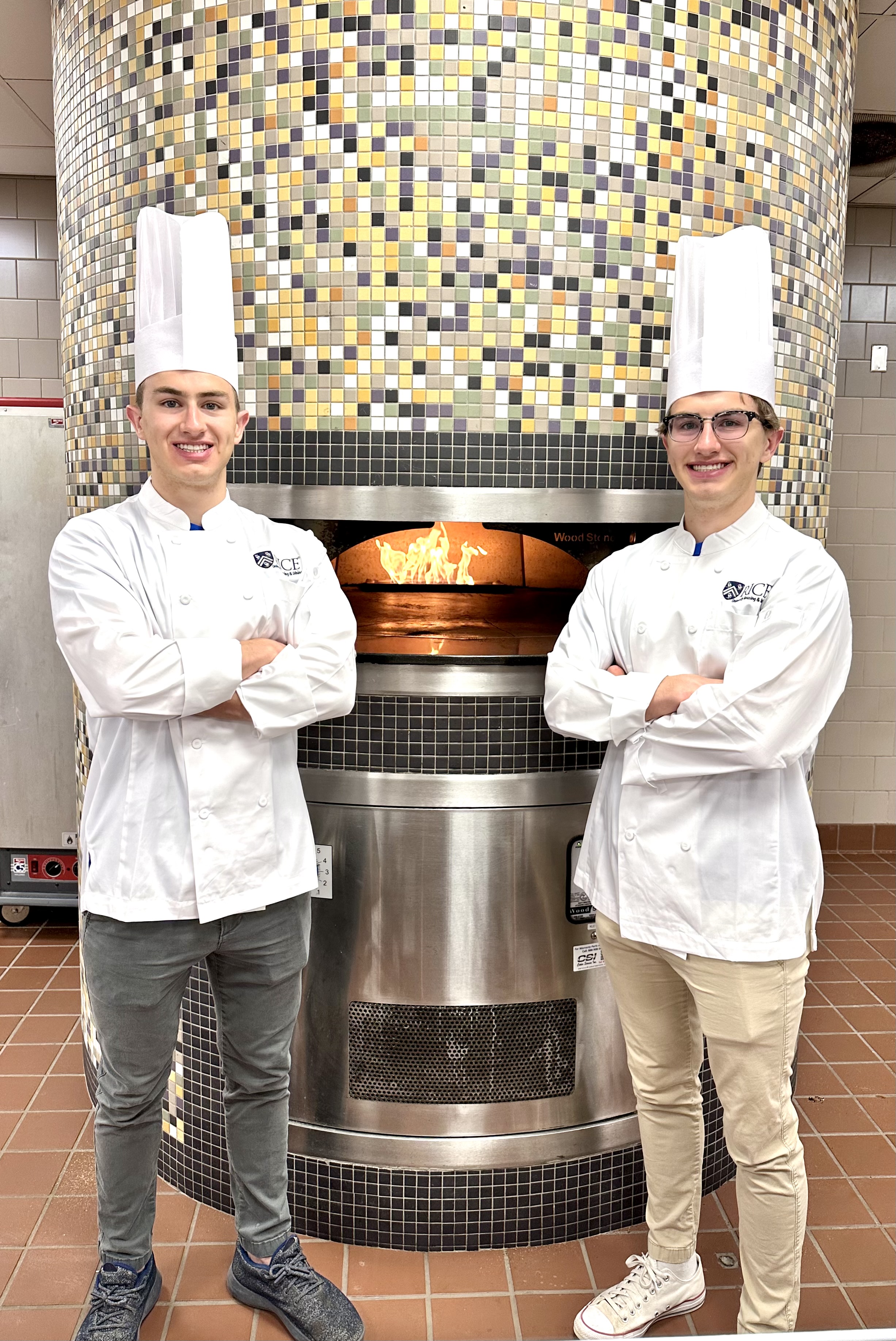 Two male students wearing a chef coat and chef hat with arms crossed in front of a pizza oven.