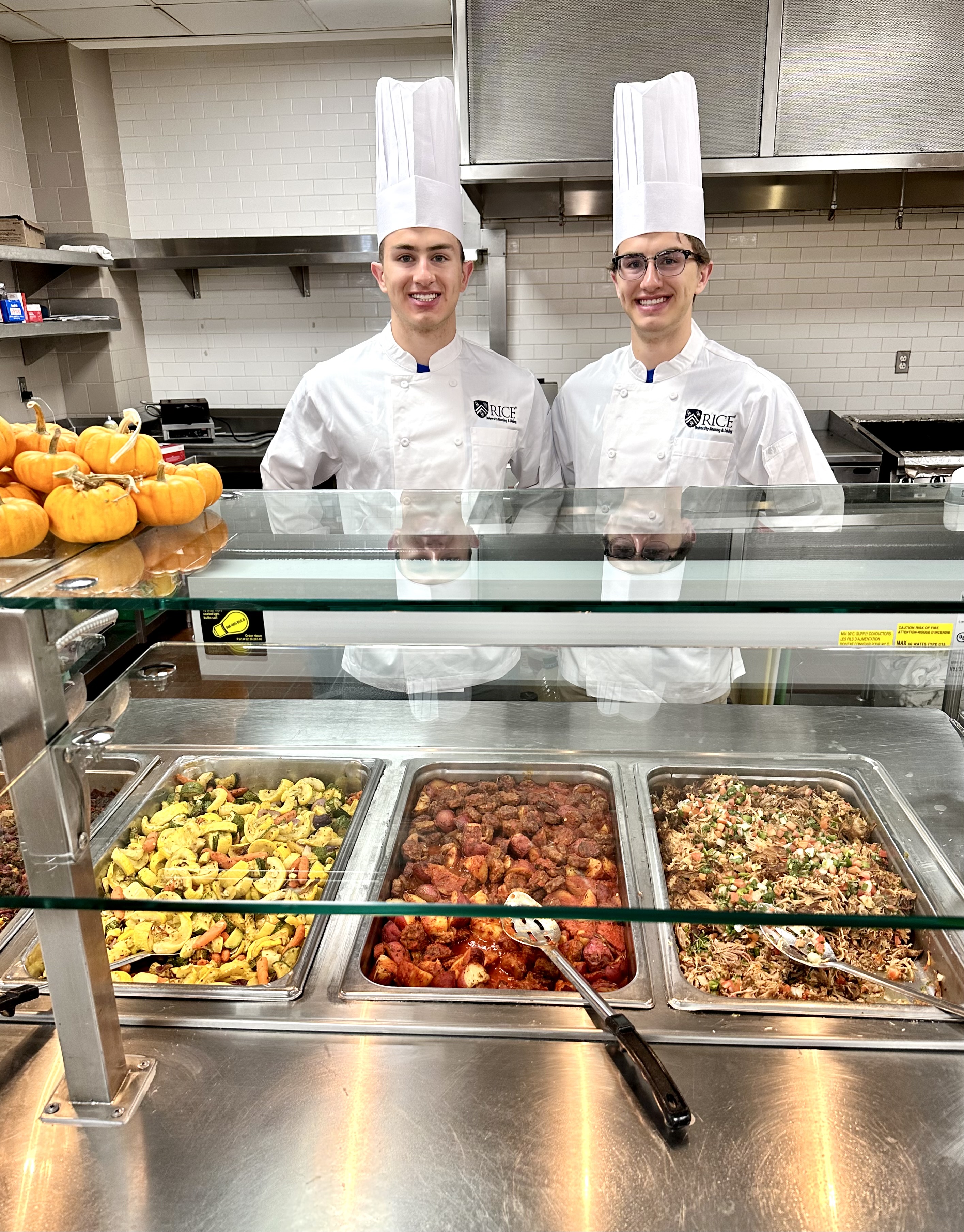 Two male students wearing a chef coat and chef hat standing in front of food counter.