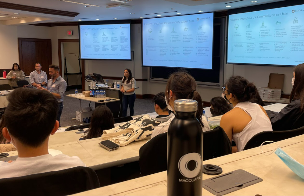 People sitting in a classroom listening to guest speakers with three projector screens behind them.