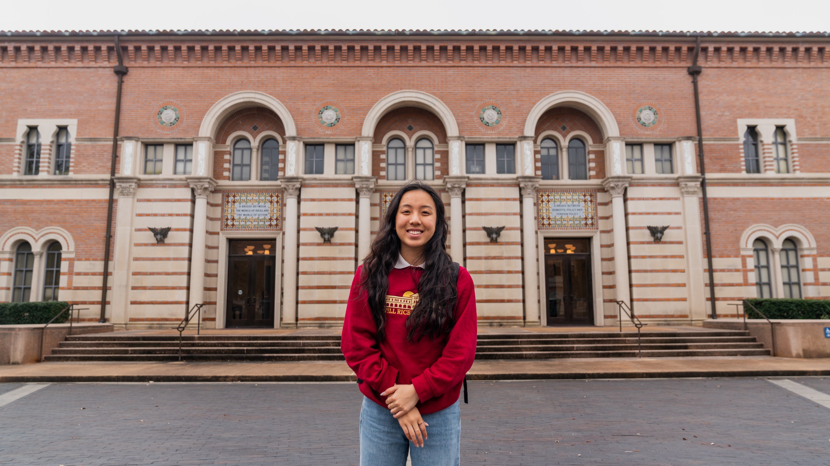 Female student wearing red crewneck standing outside of the Baker Institute, with the fountain behind her.