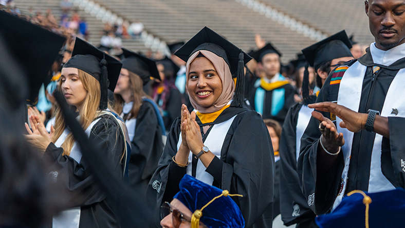 Female student wearing full graduation regalia set and head cover smiling and clapping.