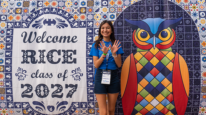Female holding up her hands in Rice owl formation in front of a tapestry mural that says "Welcome Class of 2027".