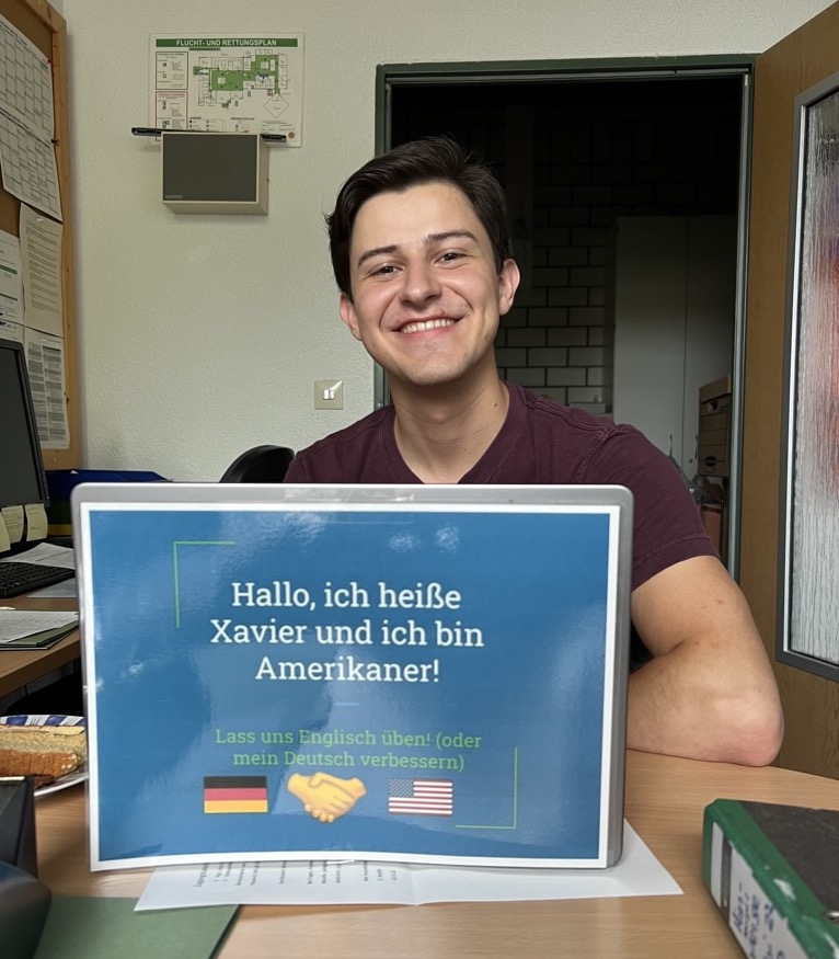 Xavier smiling behind a sign with German on it.