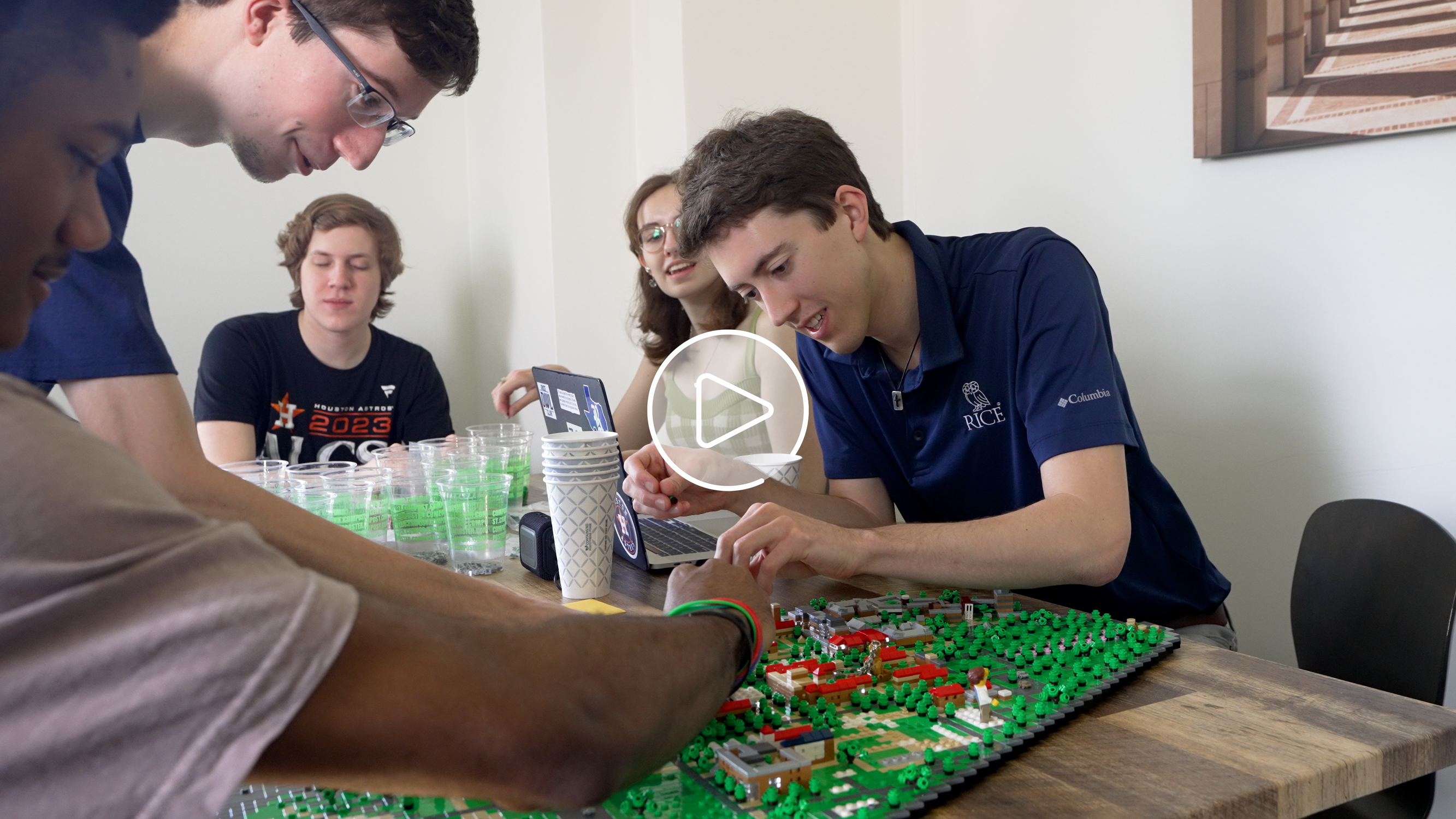 Andrew Linhart working on the LEGO Rice University build with other students with play button superimposed on top.