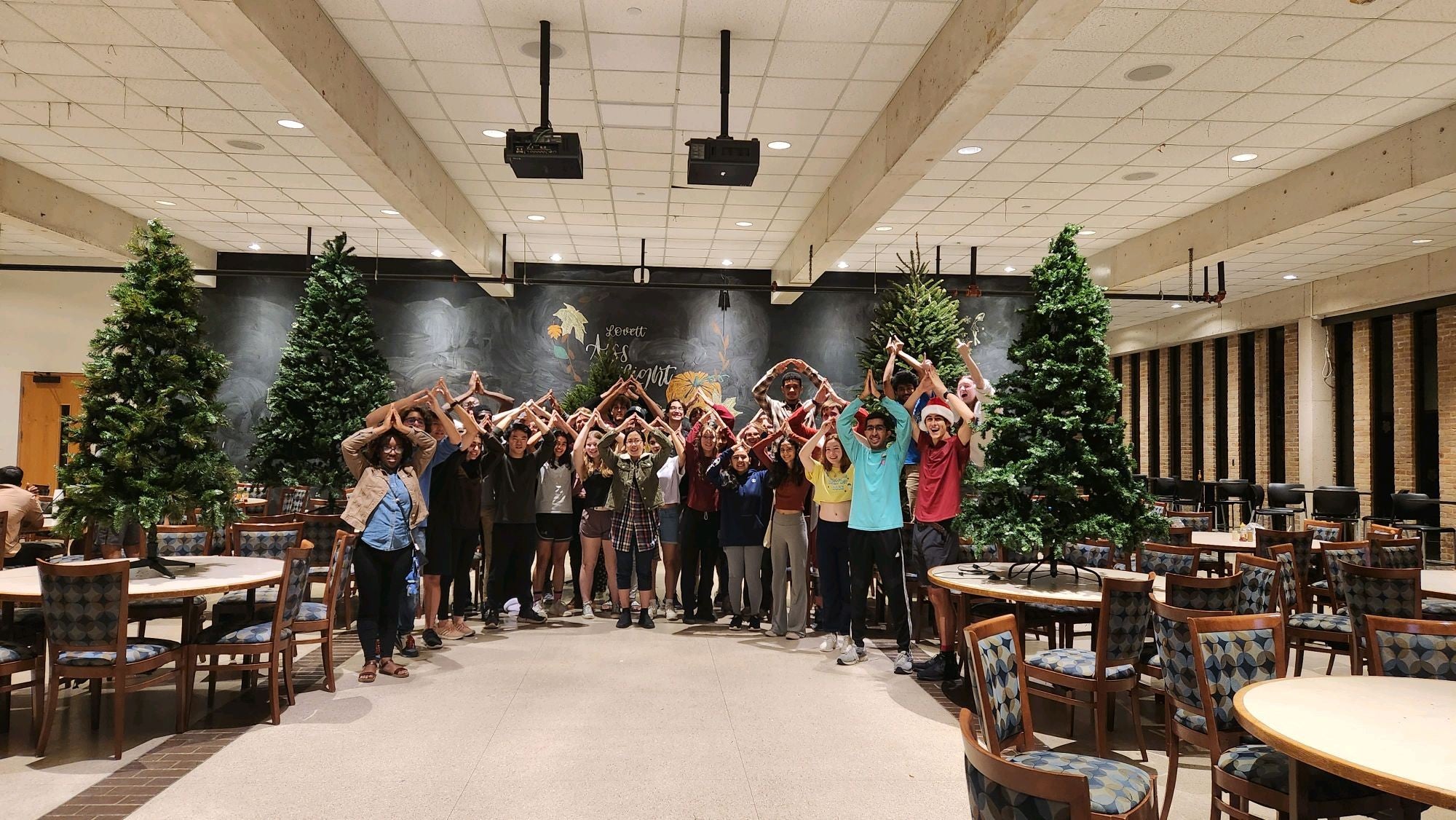 A group of college students posing in front of Christmas trees, making an A over their heads with their arms 