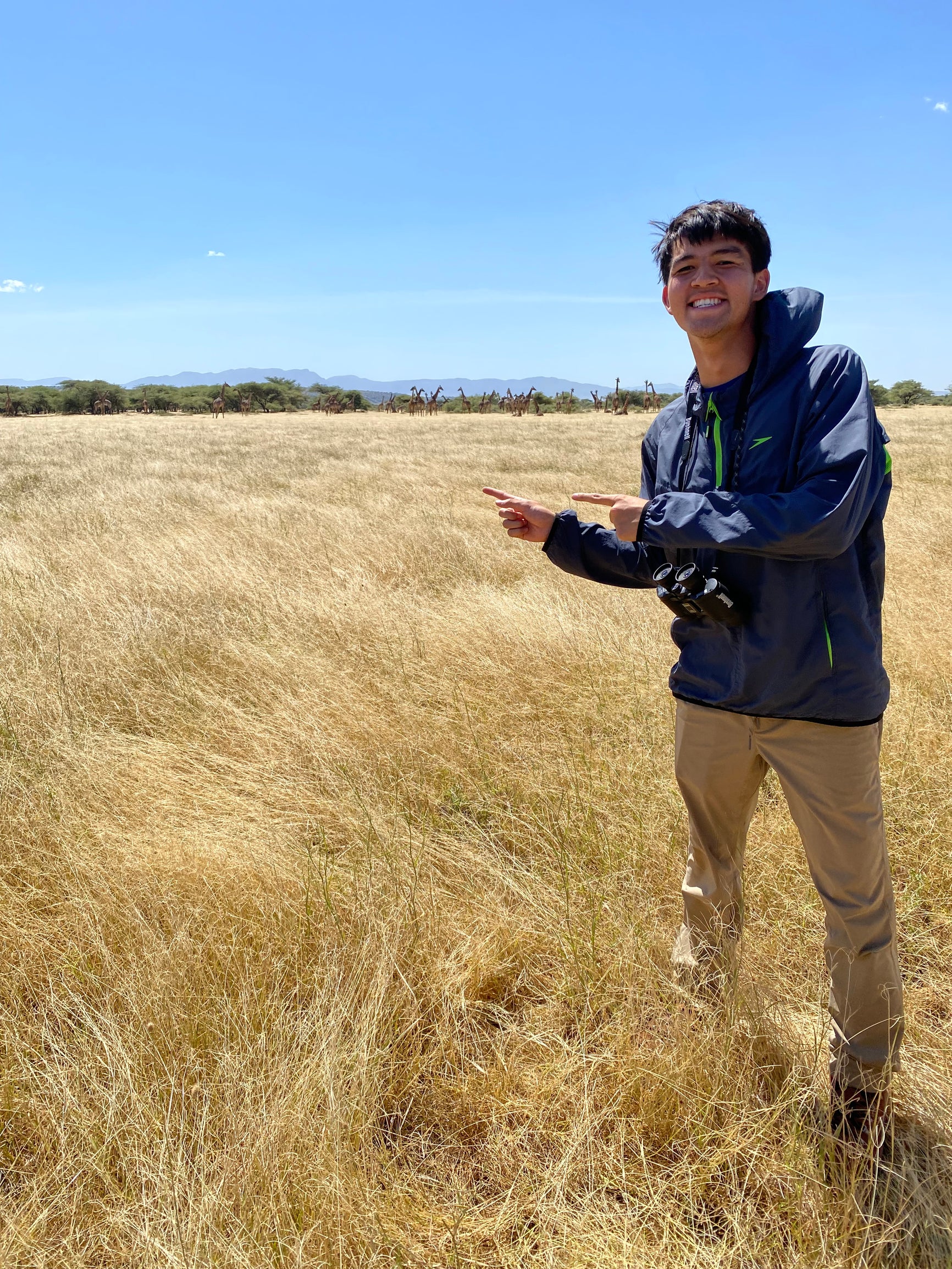 A college student wearing a sweatshirt and pants, posing in tall grass in front of a bright blue sky