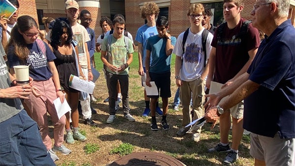 A group of students on campus, gathered around a professor as he gives a lecture.