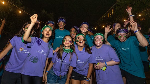 A group of students sporting matching shirts and face paint cheer together during nighttime