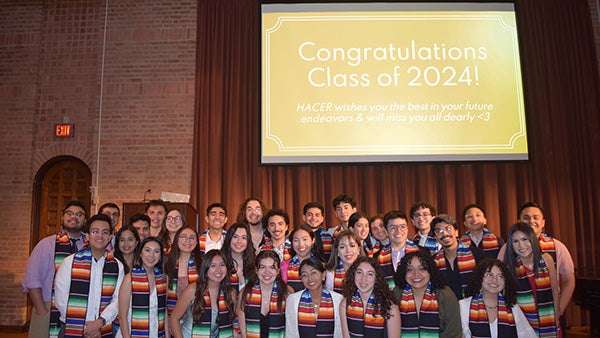 Students from cultural group HACER pose in matching stoles under a slide that says "Congrats Class of 2024".
