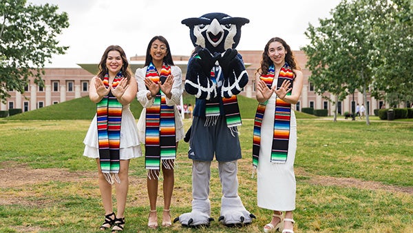 Three students in colorful stoles and the Rice Owl mascot hold out matching Rice hand signs.