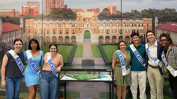 Six students in formal clothes and "Senior" sashes pose on opposite sides of a large image of Lovett Hall at Rice University.