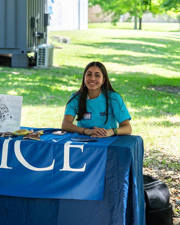 A Rice student sits at a Rice-branded table at an outdoor fair.