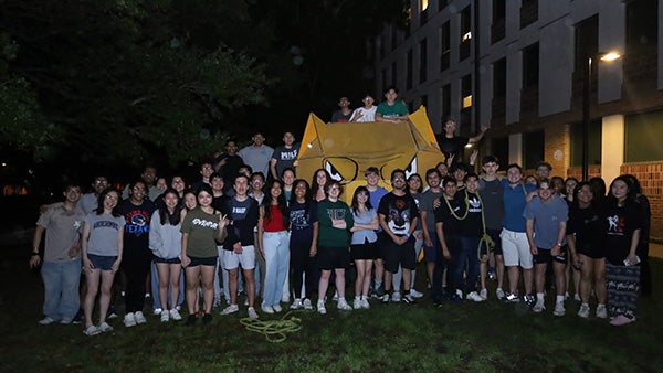 Numerous Hanszen residential college students pose with their mascot outside their building at night.