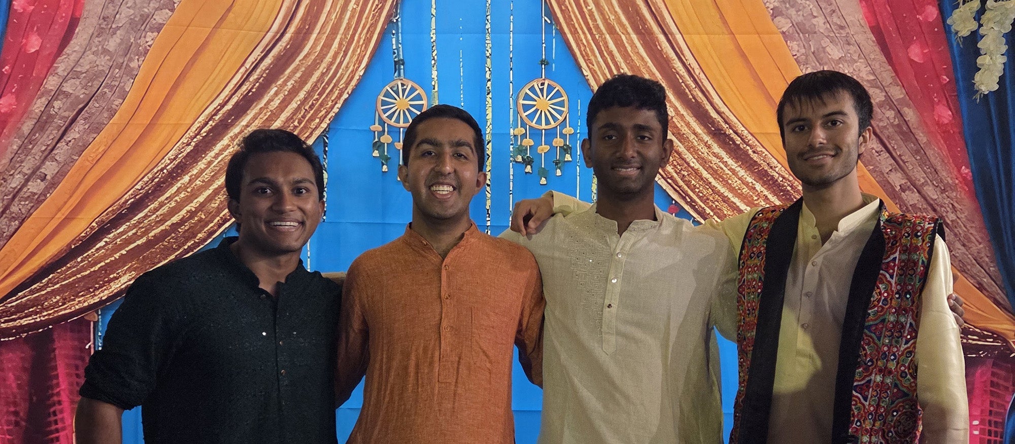 Four young people in Indian traditional clothing standing in front of a festive Indian backdrop