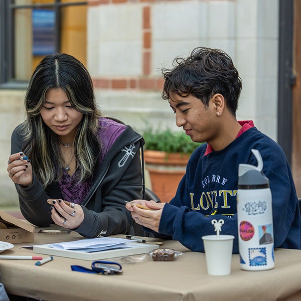 Two young people sitting at a desk outdoors, holding stationary supplies