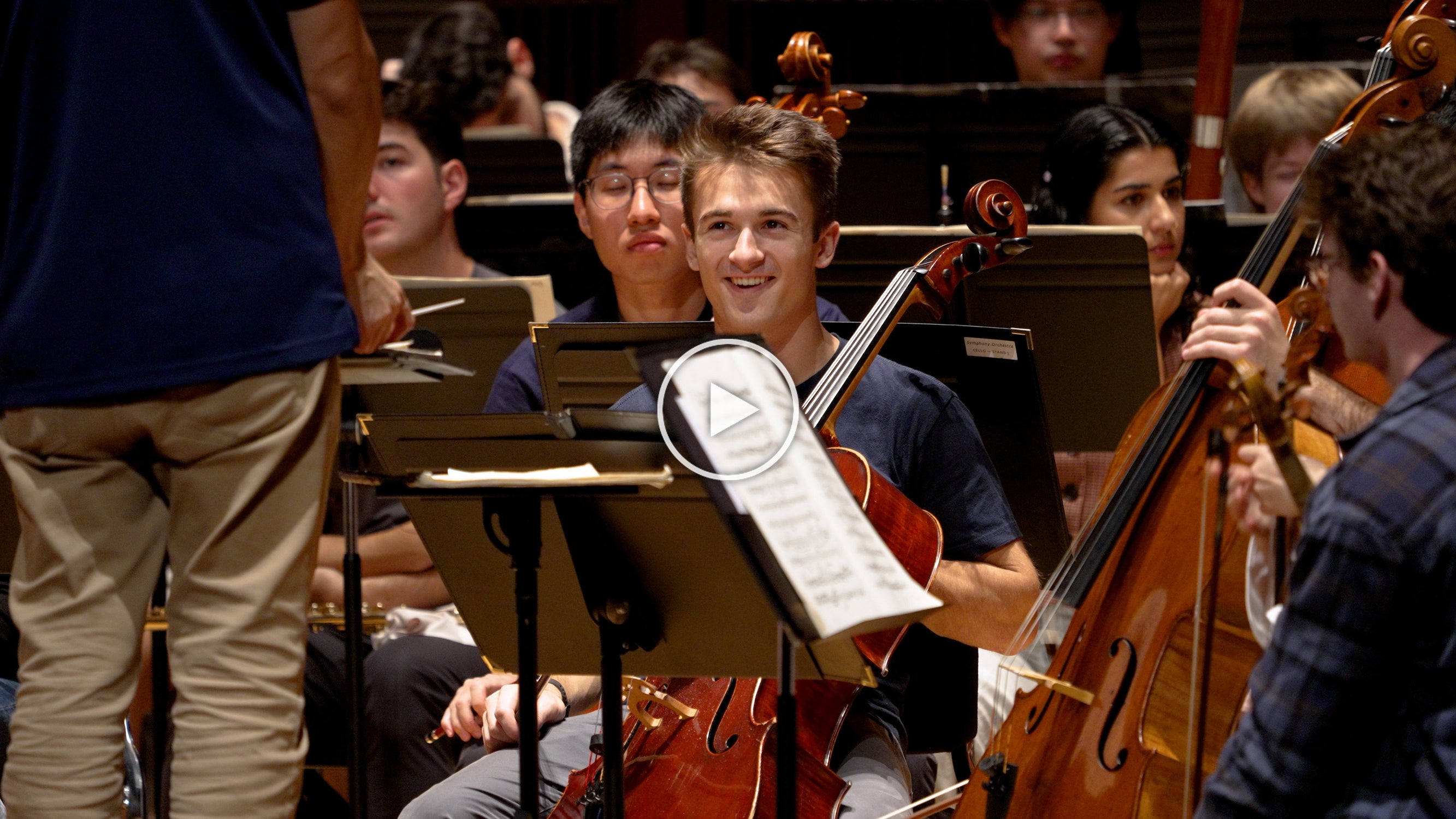 Man seated in the middle of an orchestra, looking up at a man who is partially offscreen