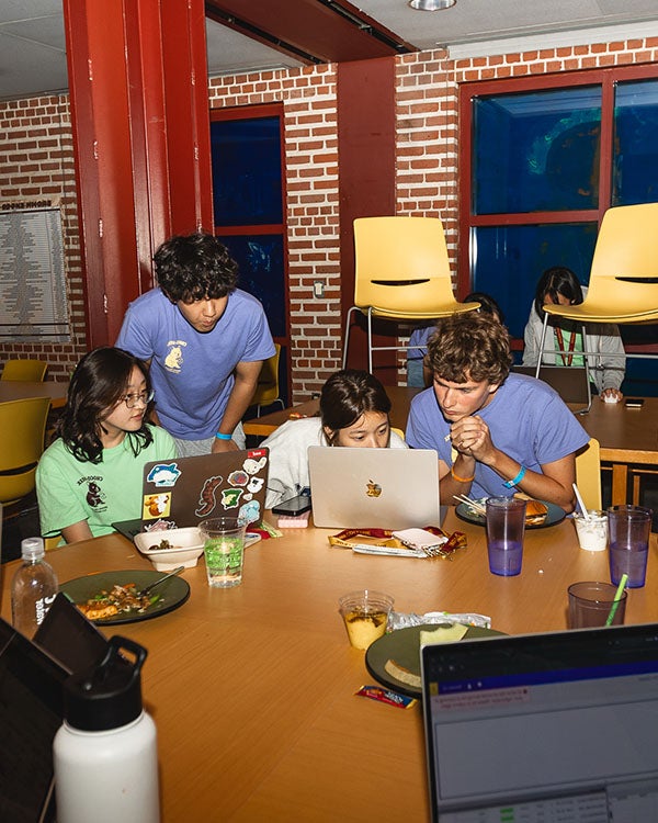 People studying around a round table, standing behind one another to look at a laptop