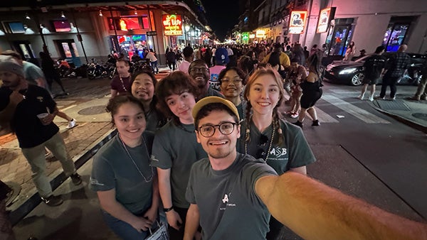 Students in matching gray shirts take a selfie on a busy street at night