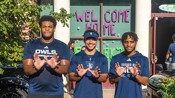 Three males in matching blue outfits, holding up their hands in owl symbols
