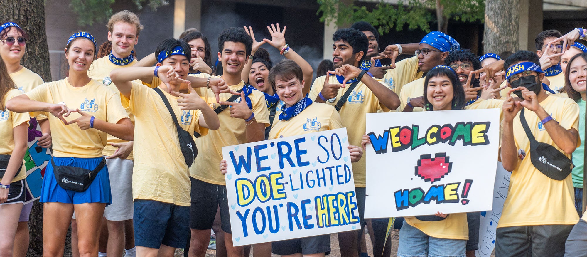 People in matching yellow shirts, two holding poster boards that say, "Welcome back" and "We're so doe-lighted you're here."