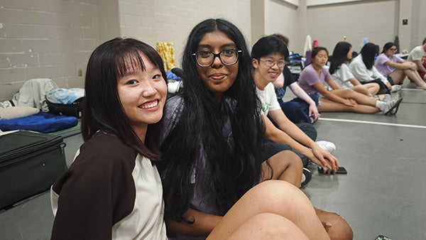 Two girls sitting, smiling at the camera, people visible behind them
