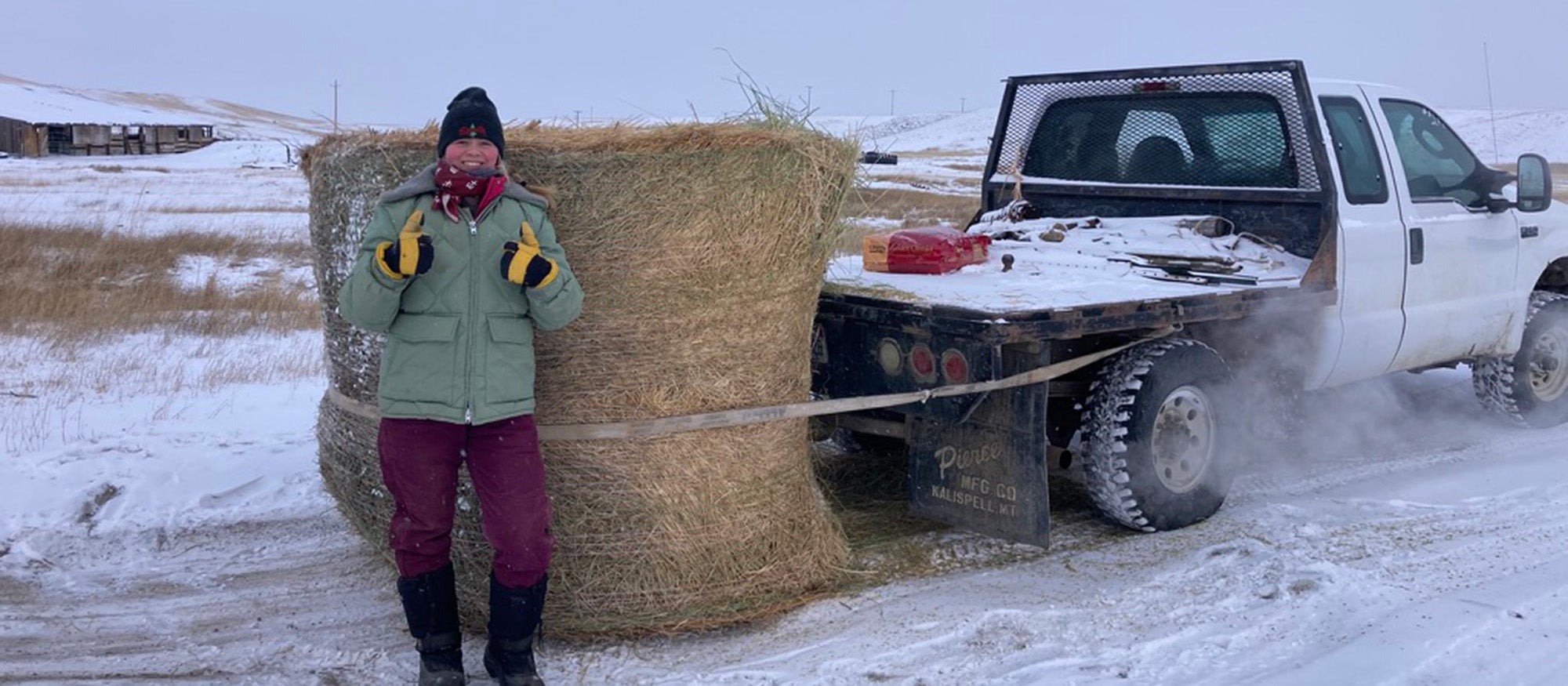 Girl in heavy winter clothing putting up two thumbs up, standing in front of large bale of hay attached to a white truck