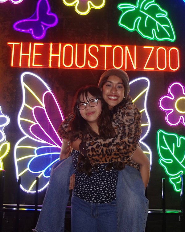 A girl on another girl's back in front of neon art displays of butterflies, flowers, leaves and word art spelling "THE HOUSTON ZOO"