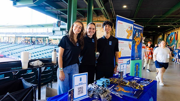 Three people standing behind a booth with various merch on it, stadium seats behind them
