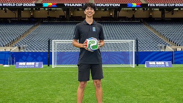 A guy holding a soccer ball, standing on a field with the goal and seats behind him; signs read "Thank you Houston", "We are Houston" and "FIFA World Cup 2026"