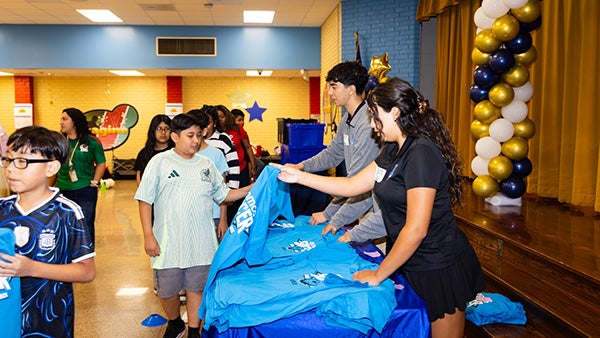 Two people handing out T-shirts to young children inside a school