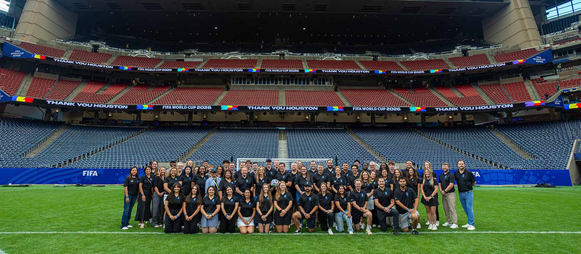 Large crowd of people standing on a soccer field in a stadium, with 3 tiers of seating visible behind them