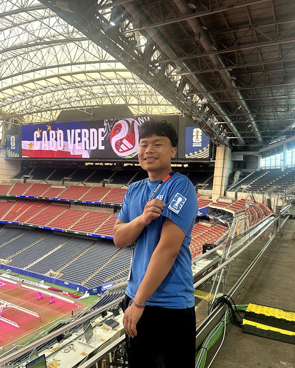 A guy gesturing to the sleeve of his shirt whilst standing on the rim of a massive stadium; a sign that says, "CABO VERDE" is visible in the background