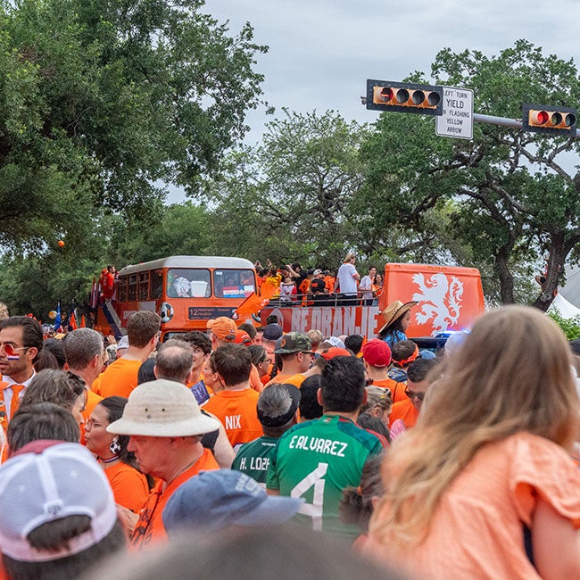 Large crowd of people following two orange buses past a traffic light and through trees