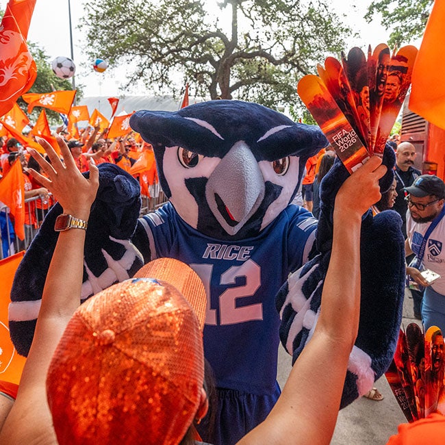 A blue Owl mascot surrounded by people in orange, walking toward a person with an orange hand fan