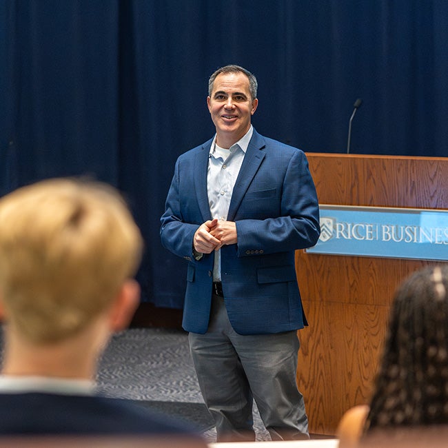 A man in a blue suit standing on a stage, speaking to partially visible students in the foreground