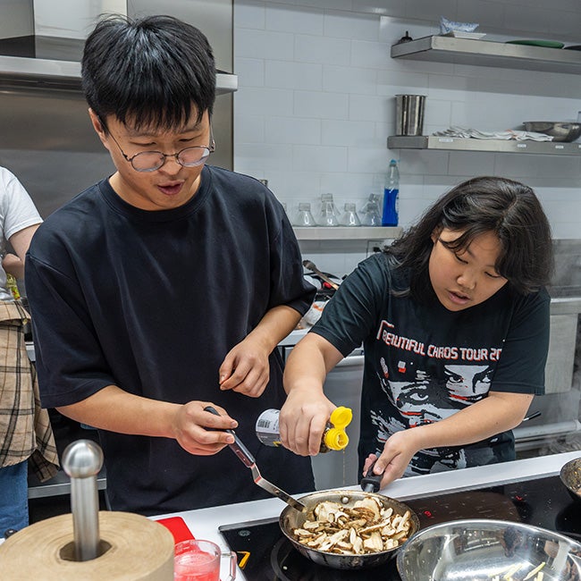 Two people cooking over a stove; one pours oil and holds the pan's handle, while the other holds a spatula