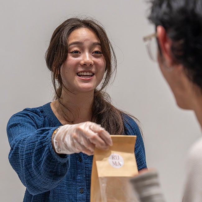 A girl wearing gloves, holding out a paper bag to a person partially visible in the shot