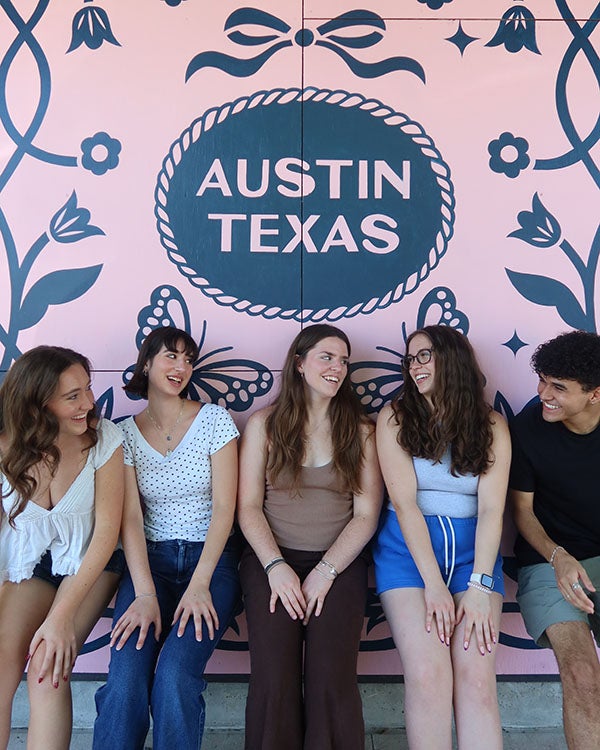 Five people laughing amongst one another in front of a wall that says "AUSTIN TEXAS"