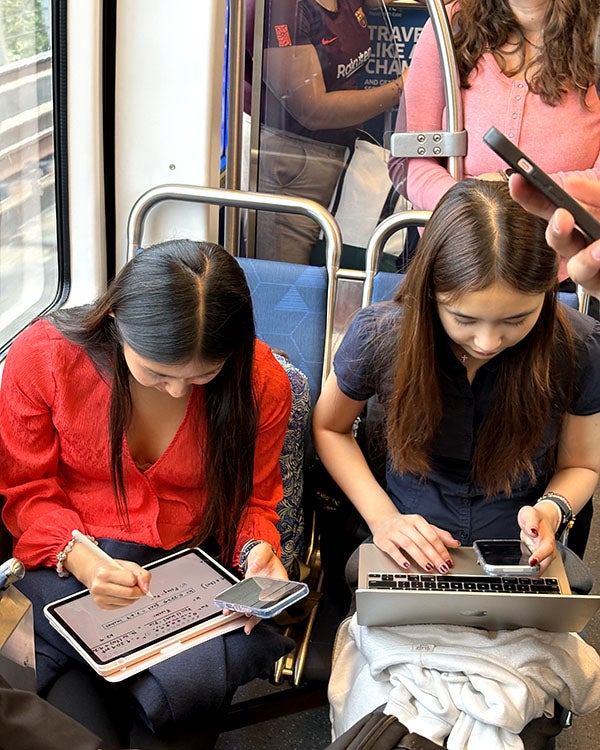 Two girls sitting on a bus, hunched over writing on their tablets