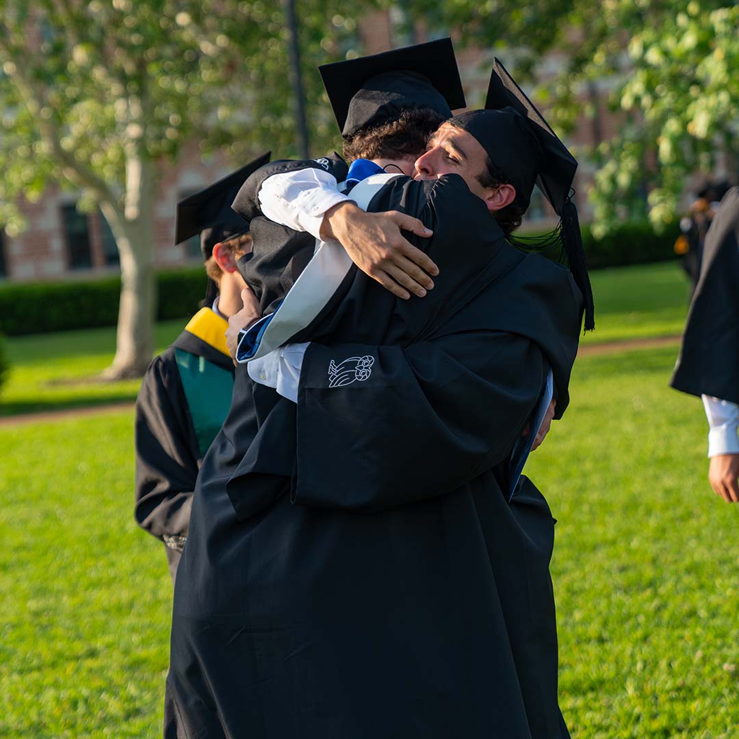 Two students wearing traditional graduation clothing share a fierce bear hug after graduation.