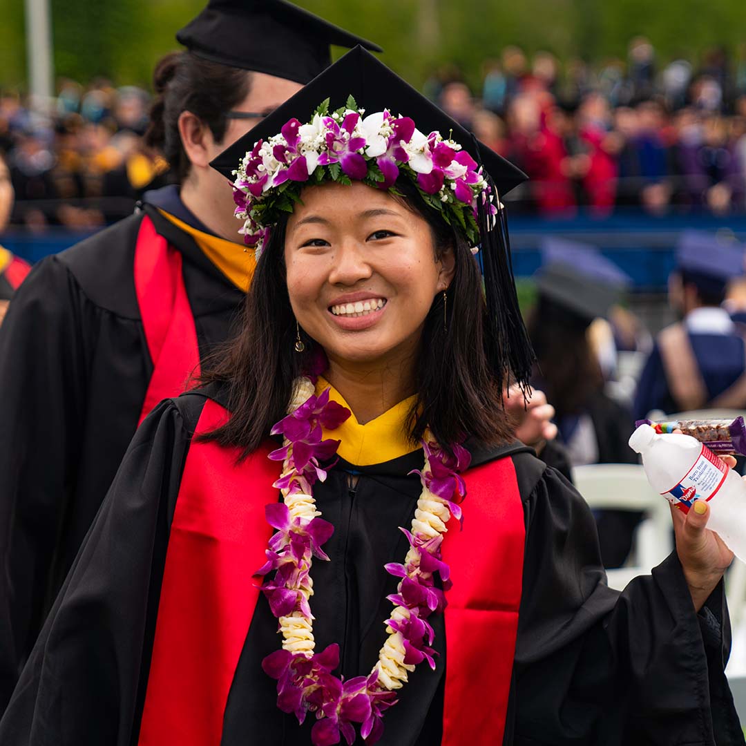 A female student wearing traditional leis as well as traditional graduation gear smiles for a picture at the graduation ceremony.